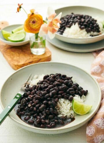 Two plates of Instant Pot black beans over rice with a lime wedge. A vase of flowers and bowl of lime wedges in-between the plates.