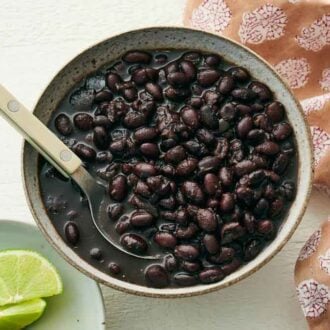 A bowl of Instant Pot black beans with a spoon. Two lime wedges on a plate and a napkin beside it.