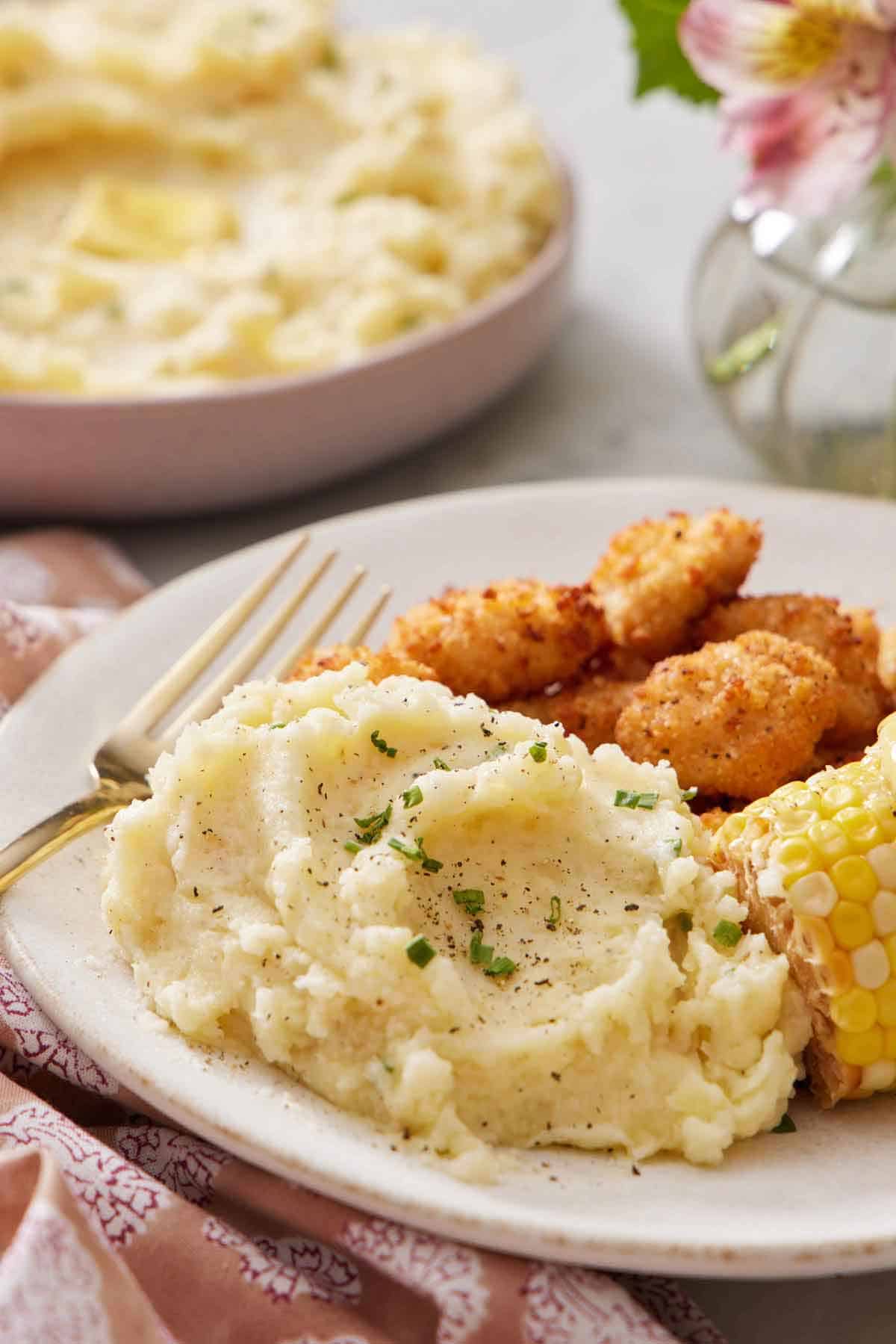 A plate of Instant Pot mashed potatoes with a fork along with some corn and breaded meat. A bowl of more mashed potatoes in the background.