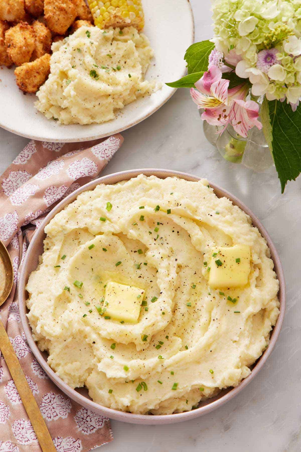 Overhead view of a bowl of Instant Pot mashed potatoes with butter, chives, and pepper. A vase of flowers and plated meal behind it.