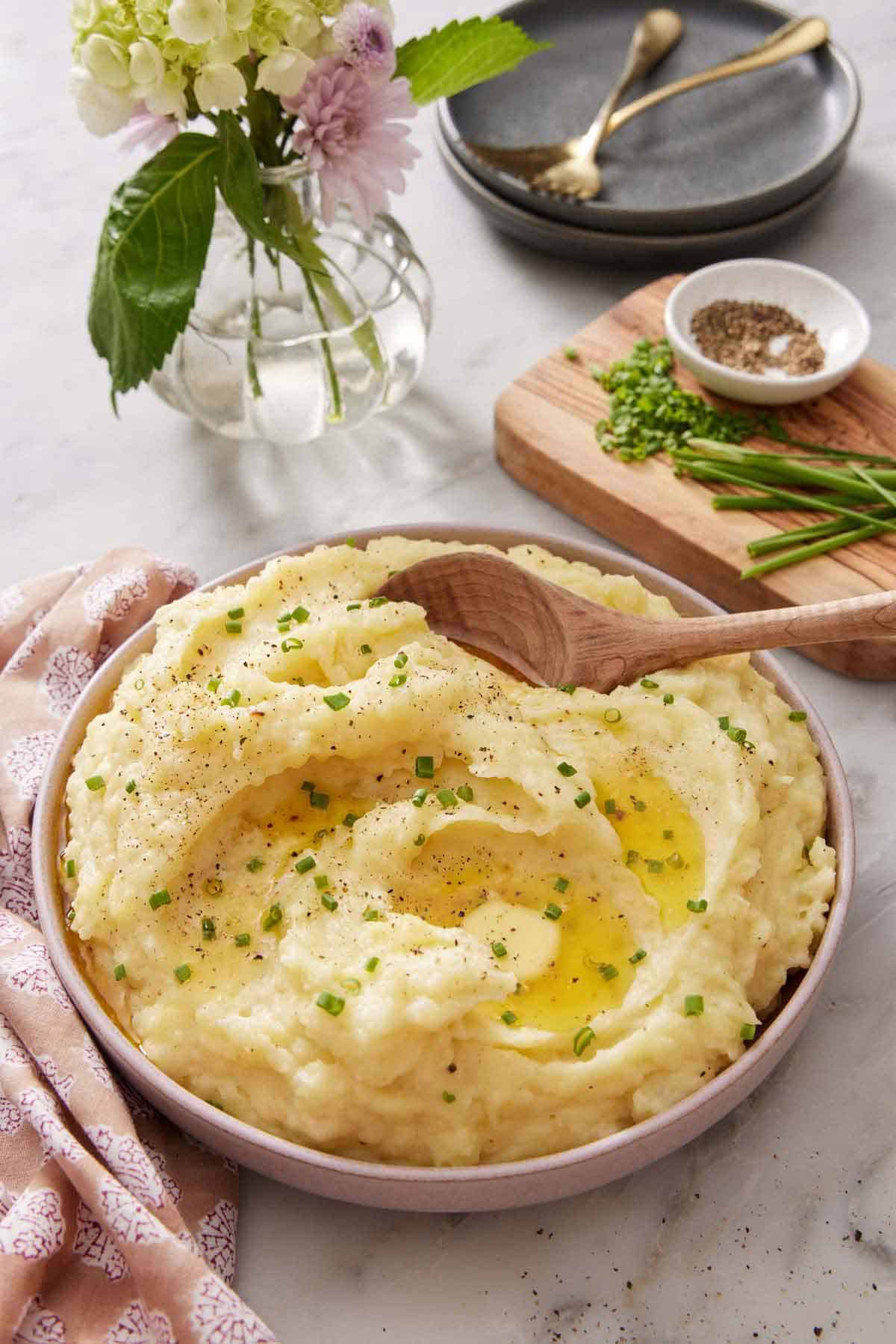 A bowl of Instant Pot mashed potatoes topped with chives and melted butter. A wooden spoon scooped in. Additional toppings and flowers in the background.