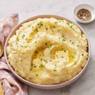 A bowl of Instant Pot mashed potatoes topped with butter, chives, and pepper. A serving spoon and a bowl of pepper beside it.