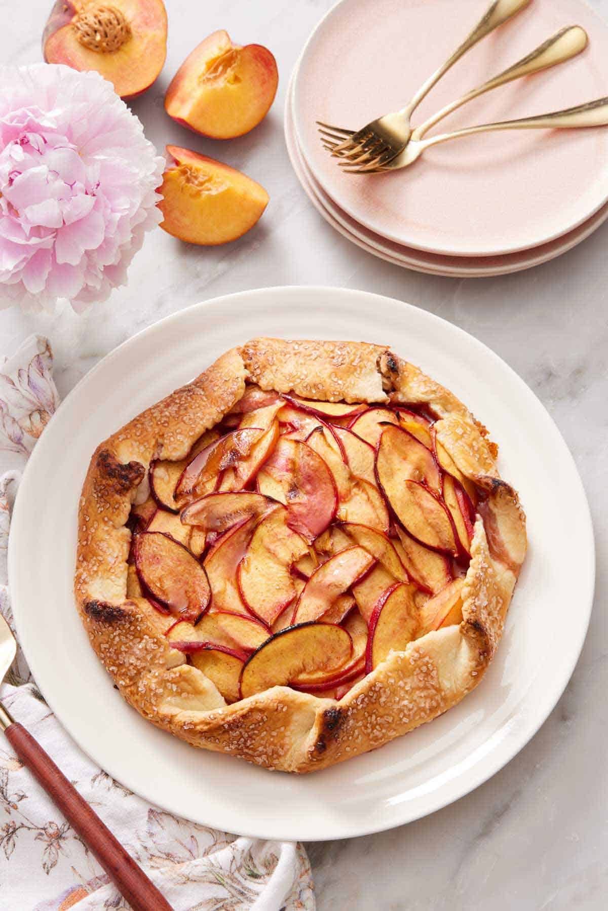 A platter with a peach galette with a stack of plates and forks in the background with peaches.