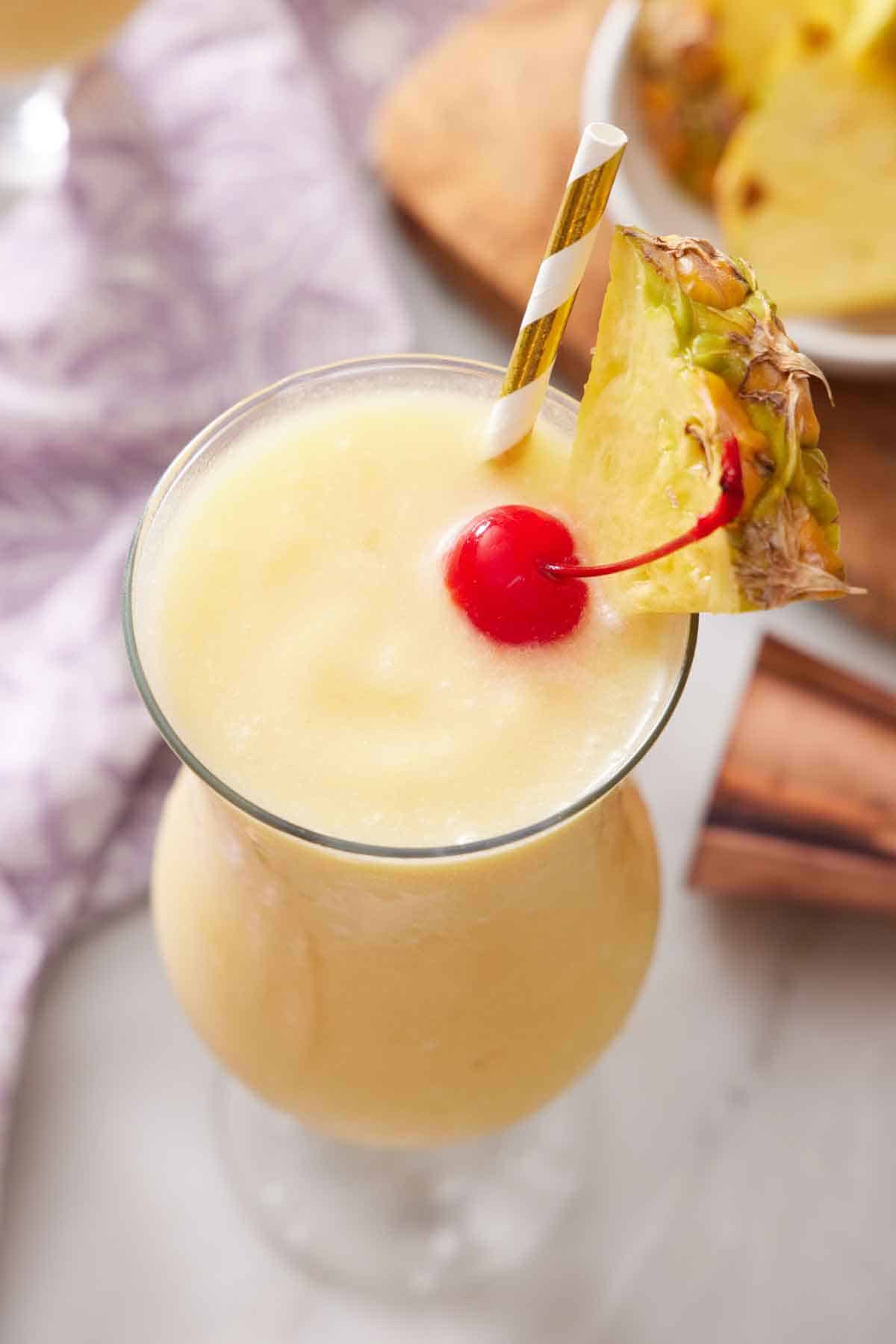 A slightly overhead view of a glass of piña colada with a straw, pineapple slice, and a maraschino cherry.