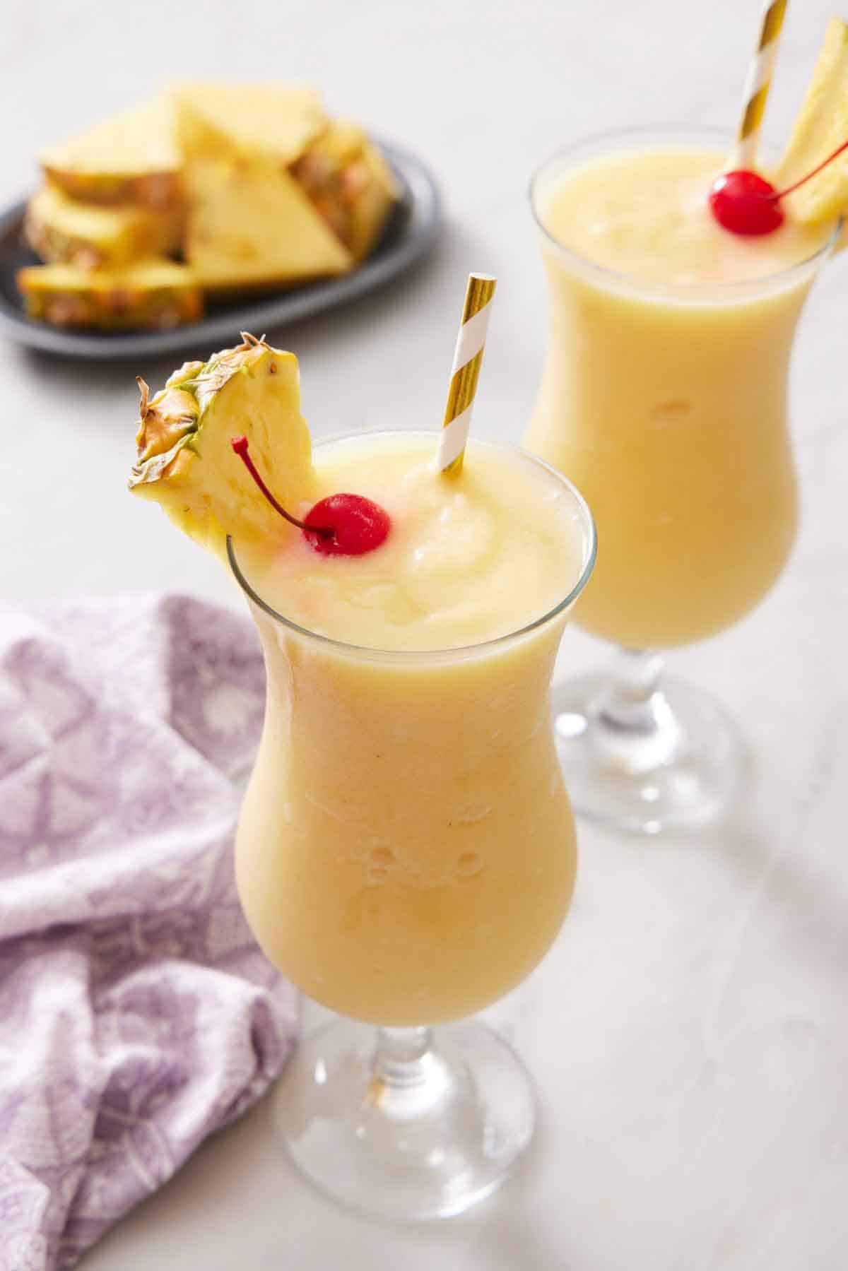 Two glasses of piña coladas, each with a straw, pineapple slice, and a maraschino cherry. Pineapple slices in the background.