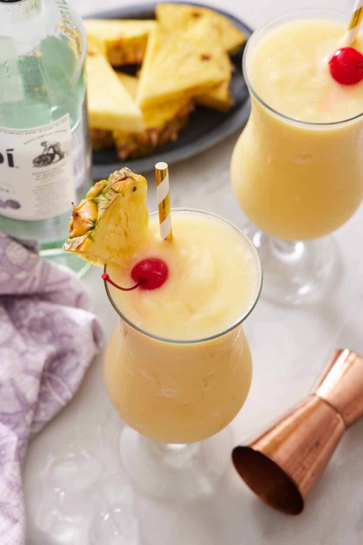 Slightly overhead view of two glasses of piña colada, each with a straw, pineapple slice, and a maraschino cherry. A jigger, platter of pineapple slices, and bottle of rum beside them.