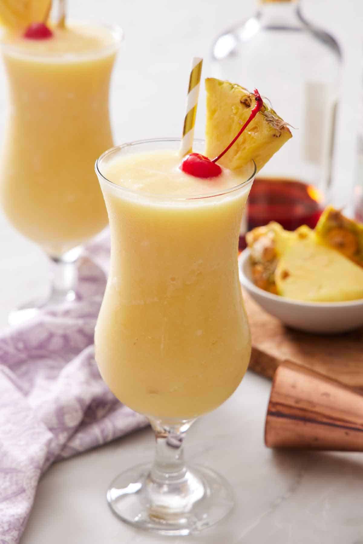 A glass of piña colada with a straw, pineapple slice, and a maraschino cherry. A second glass in the background.