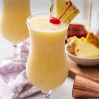 Pinterest graphic of a glass of piña colada with a straw, pineapple slice, and a maraschino cherry. A second glass in the background.