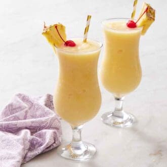 Two glasses of piña colada, each with a straw, pineapple slice, and a maraschino cherry. A linen napkin beside them.