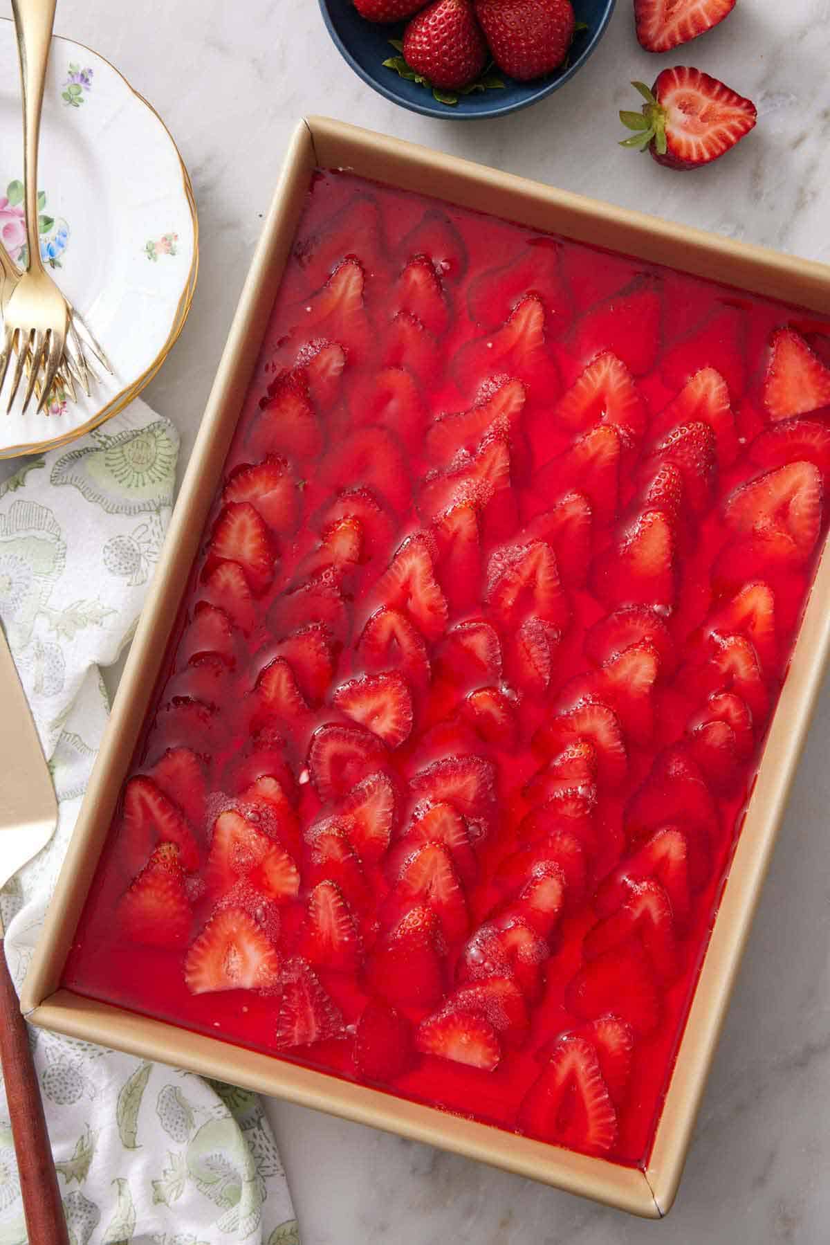 Overhead view of a rectangular baking dish of strawberry pretzel salad. Plates, forks, and strawberries on the side.