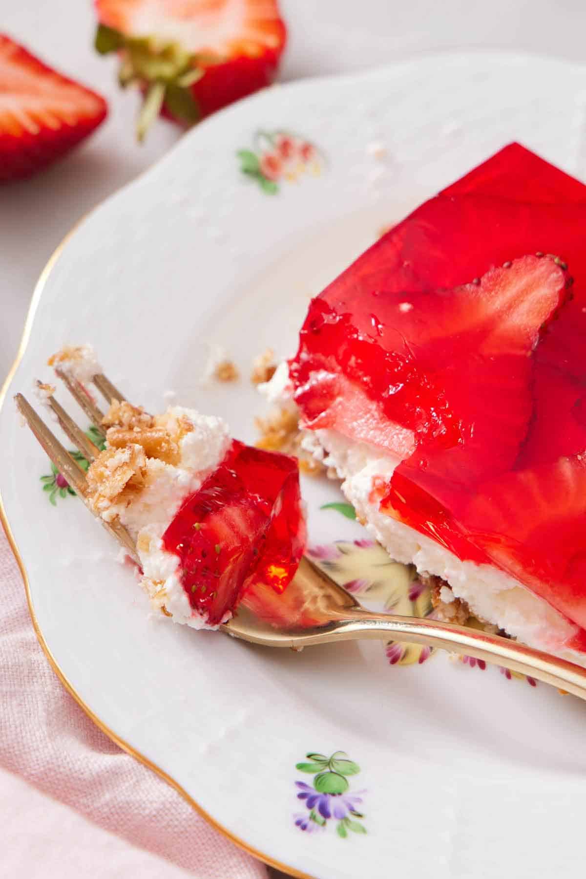 A fork with a bite of strawberry pretzel salad from a plate with a slice.