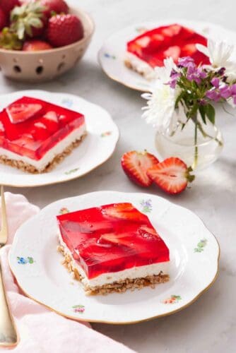 Three plates with sliced square servings of strawberry pretzel salad with a vase of flowers and bowl of strawberries between them.