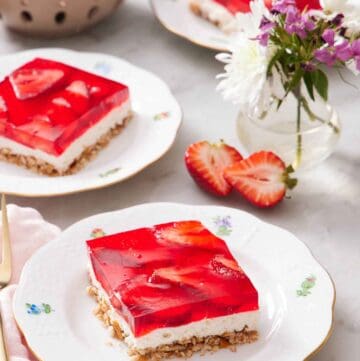 Three plates with sliced square servings of strawberry pretzel salad with a vase of flowers and bowl of strawberries between them.