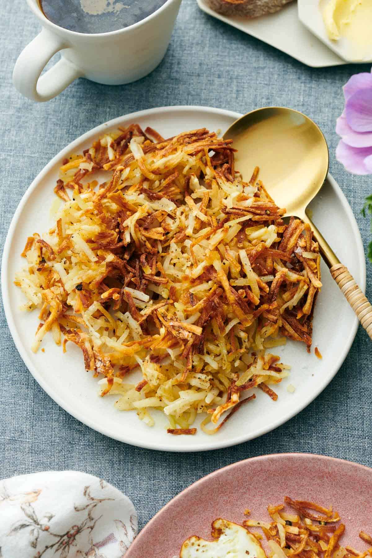 A plate of air fryer hash browns with a serving spoon. A mug of coffee behind it.