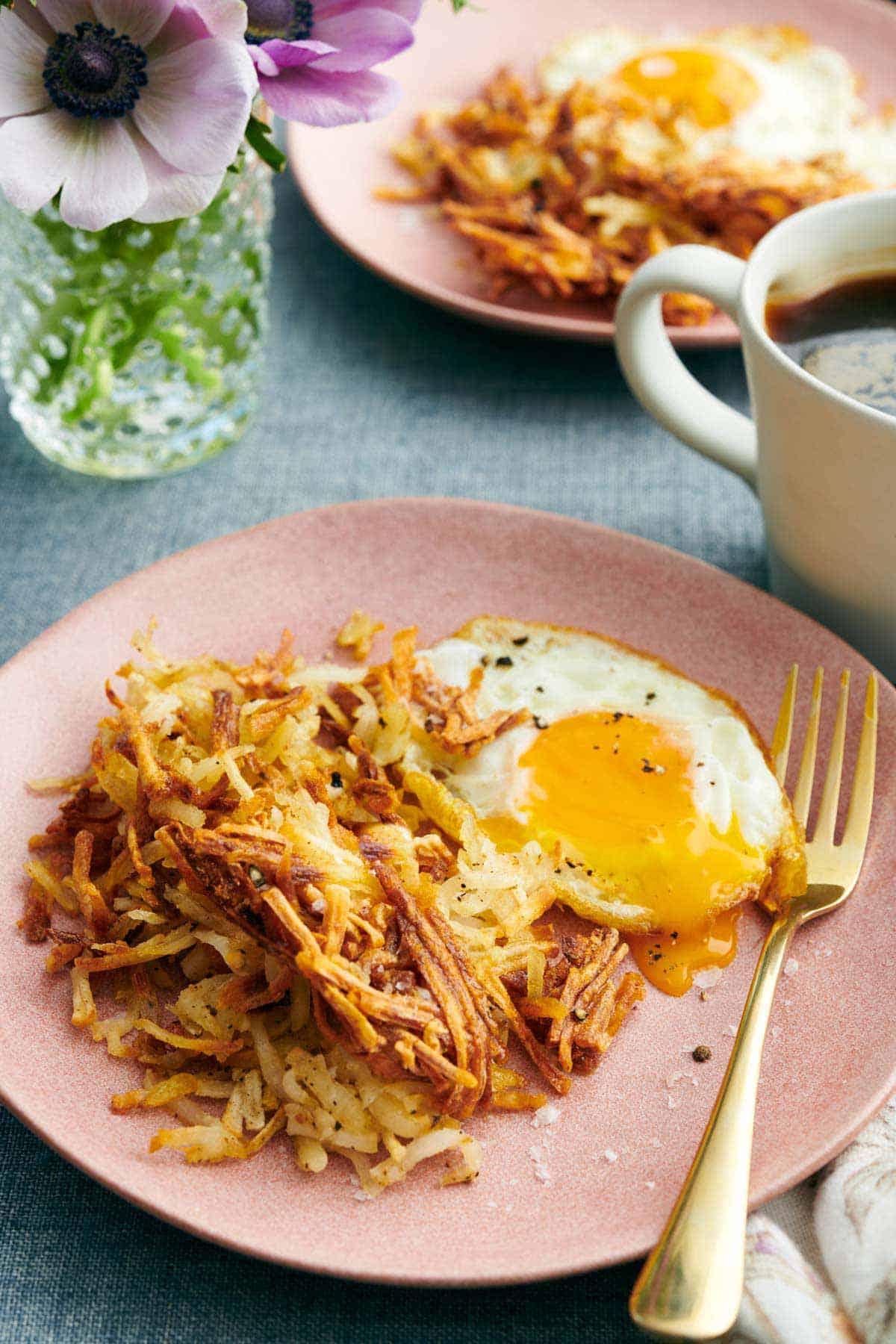 A plate of air fryer hash browns with a fried egg and a fork. A mug of coffee and another plate in the background.