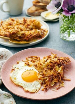 A plate with air fryer hash browns and a fried egg. A plate with more hash browns in the background along with flowers and toast.