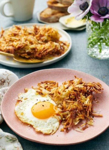 A plate with air fryer hash browns and a fried egg. A plate with more hash browns in the background along with flowers and toast.