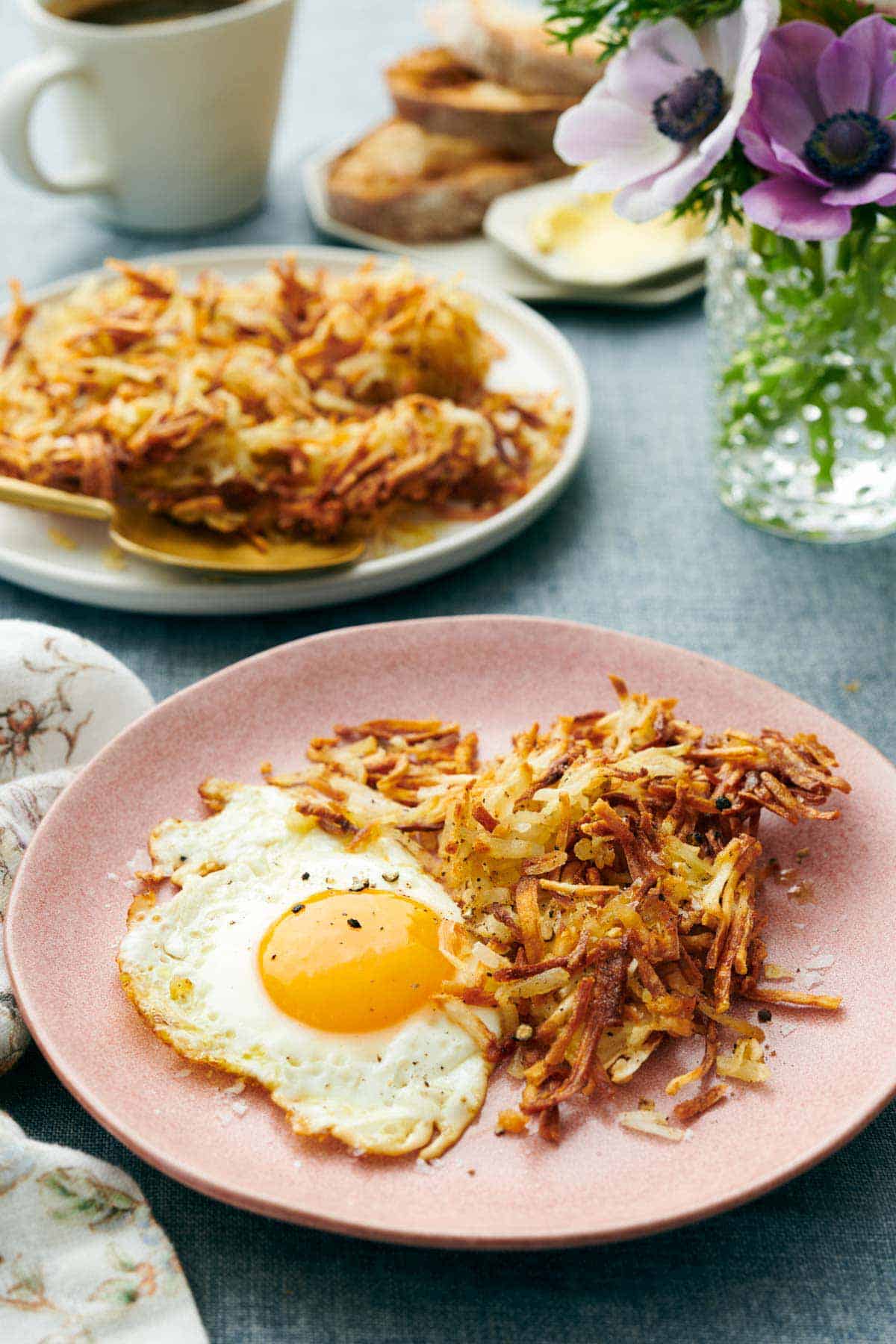 A plate with air fryer hash browns and a fried egg. A plate with more hash browns in the background along with flowers and toast.