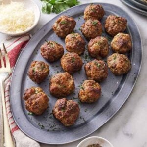 A platter with air fryer meatballs with a fork on the side. A bowl of shredded parmesan in the back.