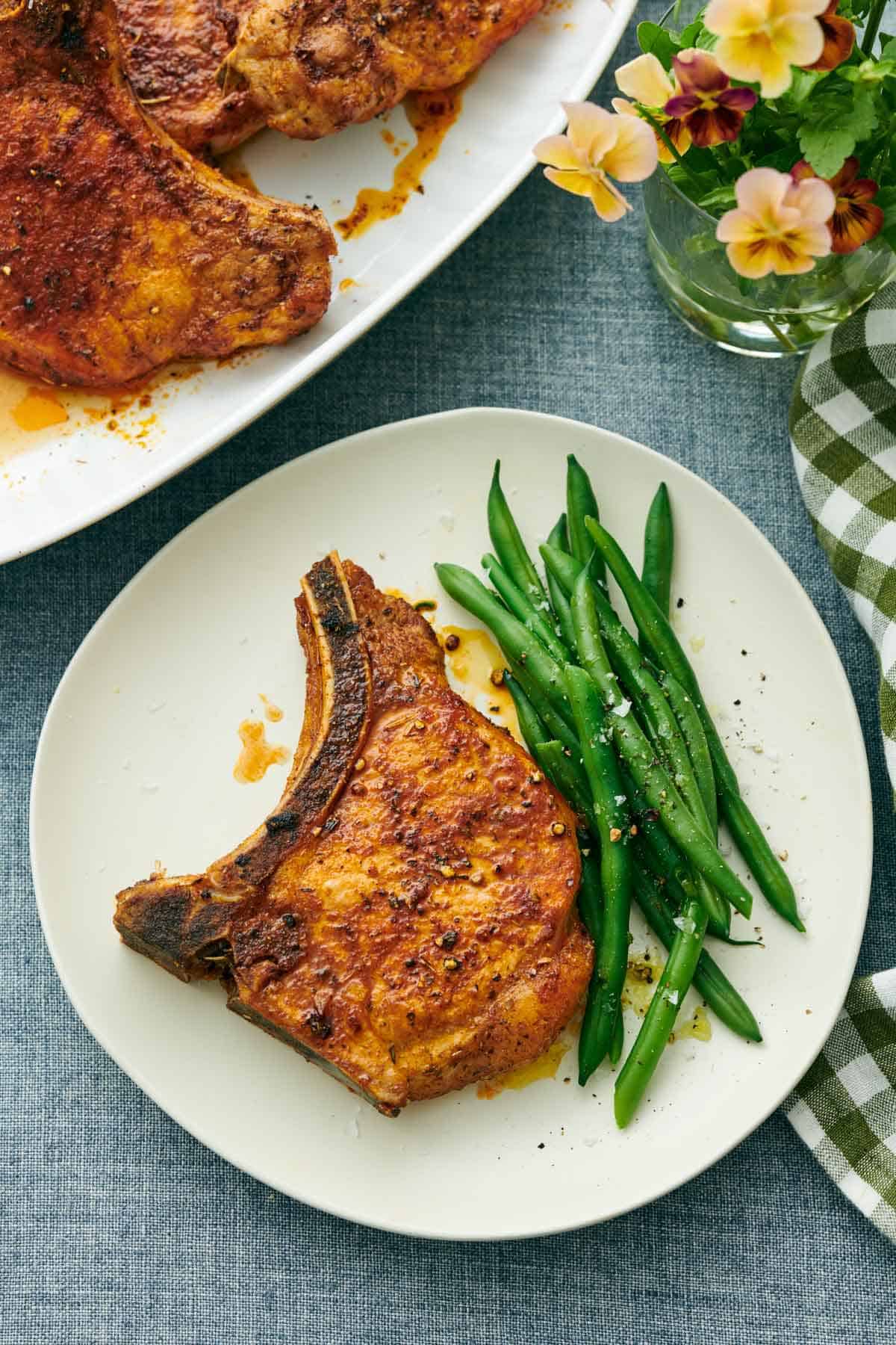 A plate with an air fryer pork chop and green beans. A platter with more pork chops and some flowers off to the side.