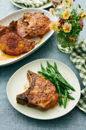 A plate with a juicy air fryer pork chop with green beans. A glass of flowers and platter of more pork chops in the background.