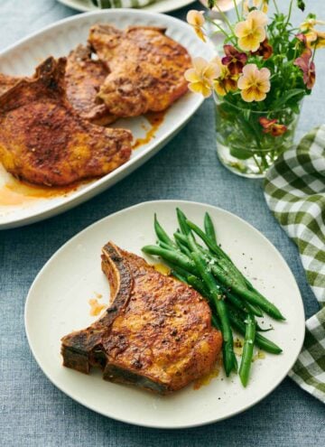 A plate with a juicy air fryer pork chop with green beans. A glass of flowers and platter of more pork chops in the background.