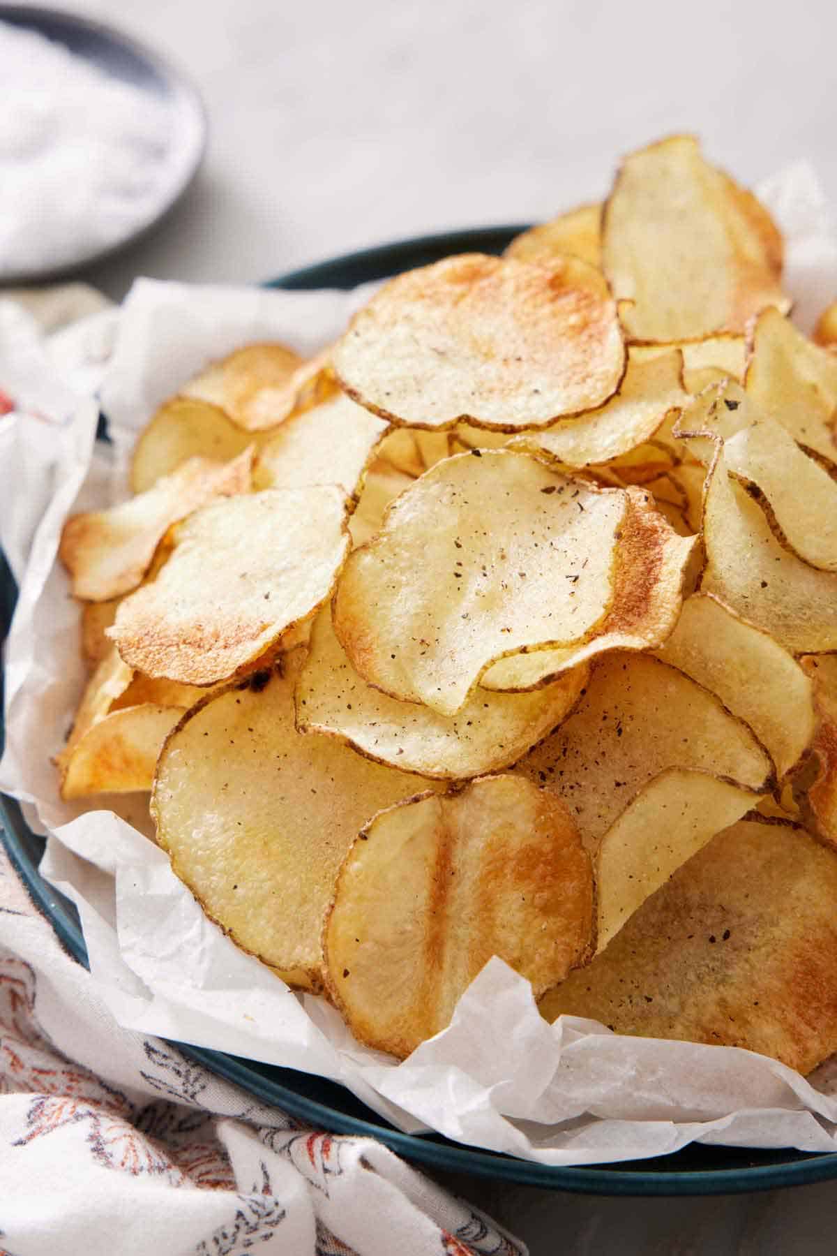 A pile of air fryer potato chips topped with some pepper in a parchment lined plate. A bowl of salt in the background.