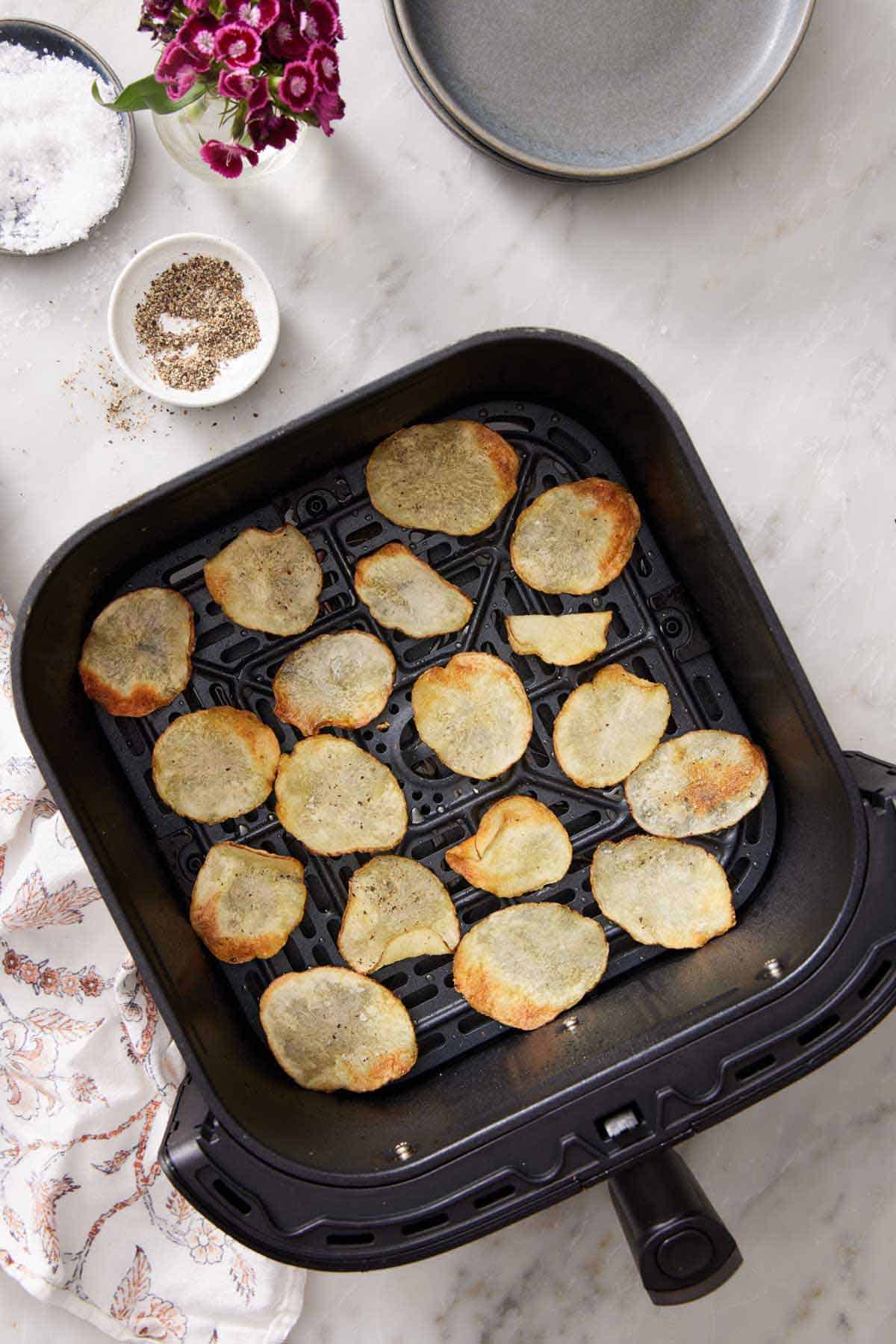 Overhead view of air fryer potato chips in an air fryer basket. A bowl of pepper, salt, some plates, and flowers off to the side.