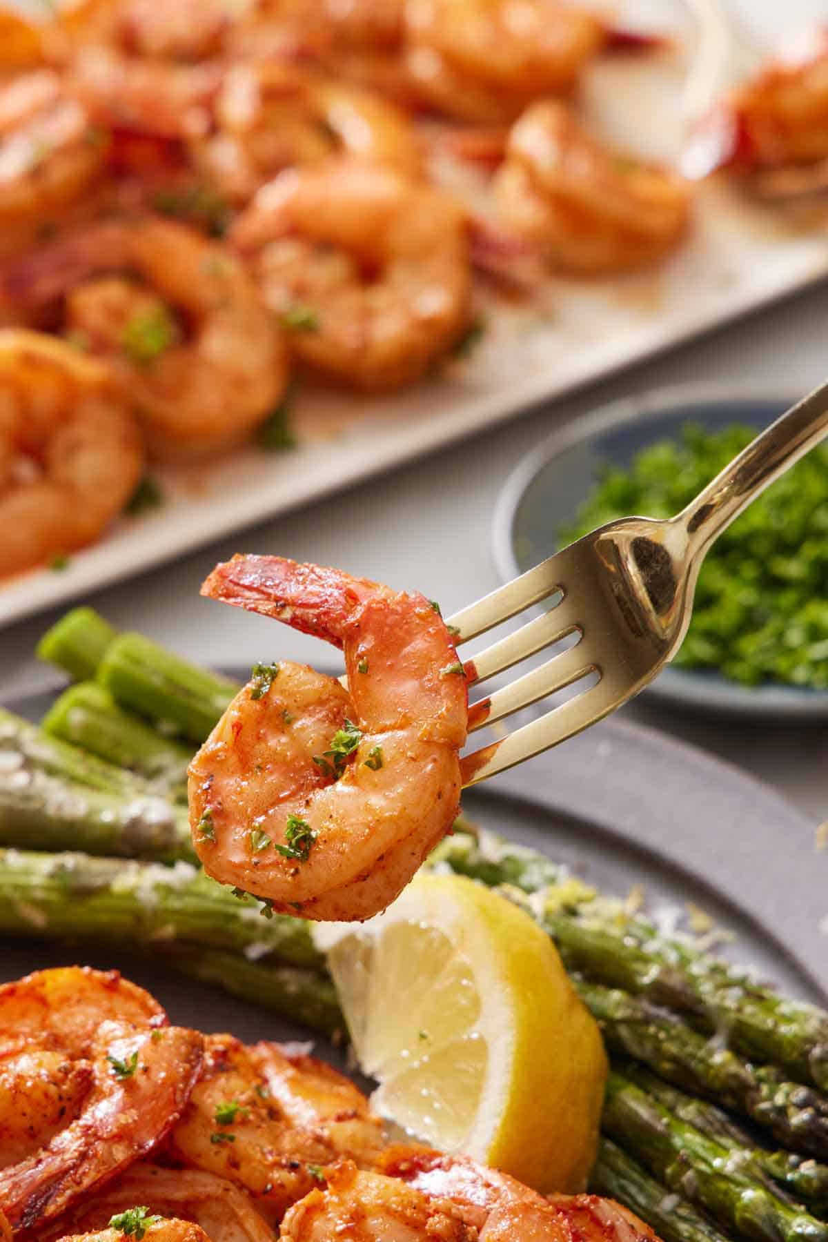 A fork with an air fryer shrimp on the end, lifted from a plate with more shrimp, lemon slices, and asparagus.