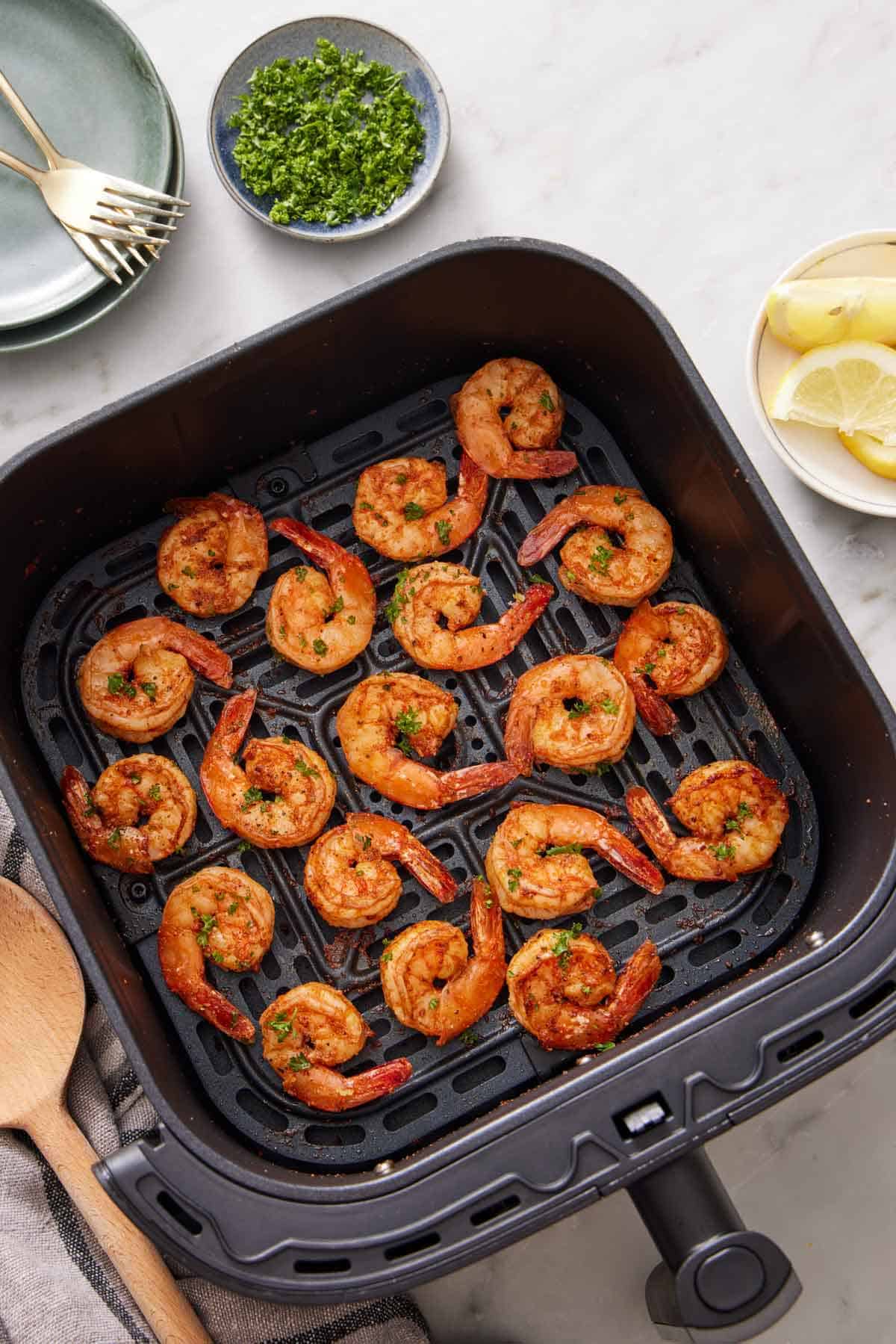 Overhead view of an air fryer basket of air fryer shrimp. A bowl of chopped parsley, lemon slices, stack of plates and forks on the side.