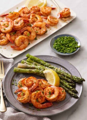 A plate with air fryer shrimps, lemon wedge, and asparagus. A platter with more shrimp in the background along with a bowl of chopped parsley.