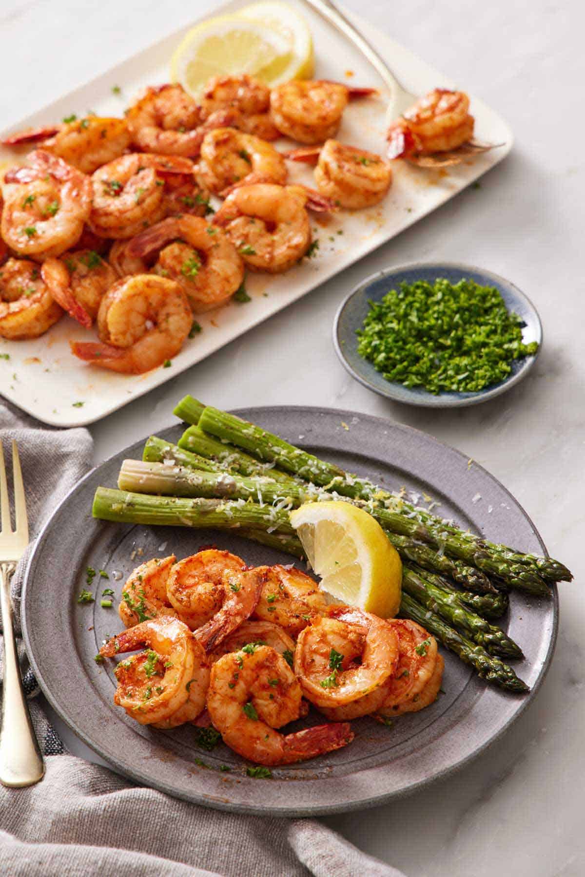 A plate with air fryer shrimps, lemon wedge, and asparagus. A platter with more shrimp in the background along with a bowl of chopped parsley.