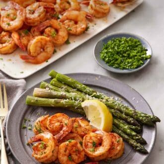 Pinterest graphic of a plate with air fryer shrimps, lemon wedge, and asparagus. A platter with more shrimp in the background along with a bowl of parsley.