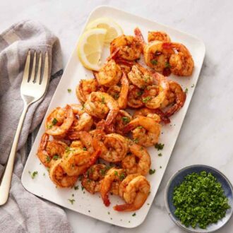 Overhead view of a platter of air fryer shrimp with lemon slices. A fork and bowl of chopped parsley on the side.