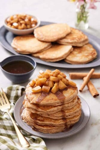 A stack of apple pancakes topped with apples and syrup. More pancakes and cooked apples in the background along with cinnamon sticks and syrup.