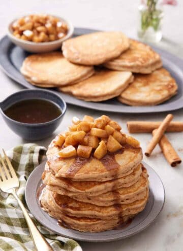 A stack of apple pancakes topped with apples and syrup. More pancakes and cooked apples in the background along with cinnamon sticks and syrup.