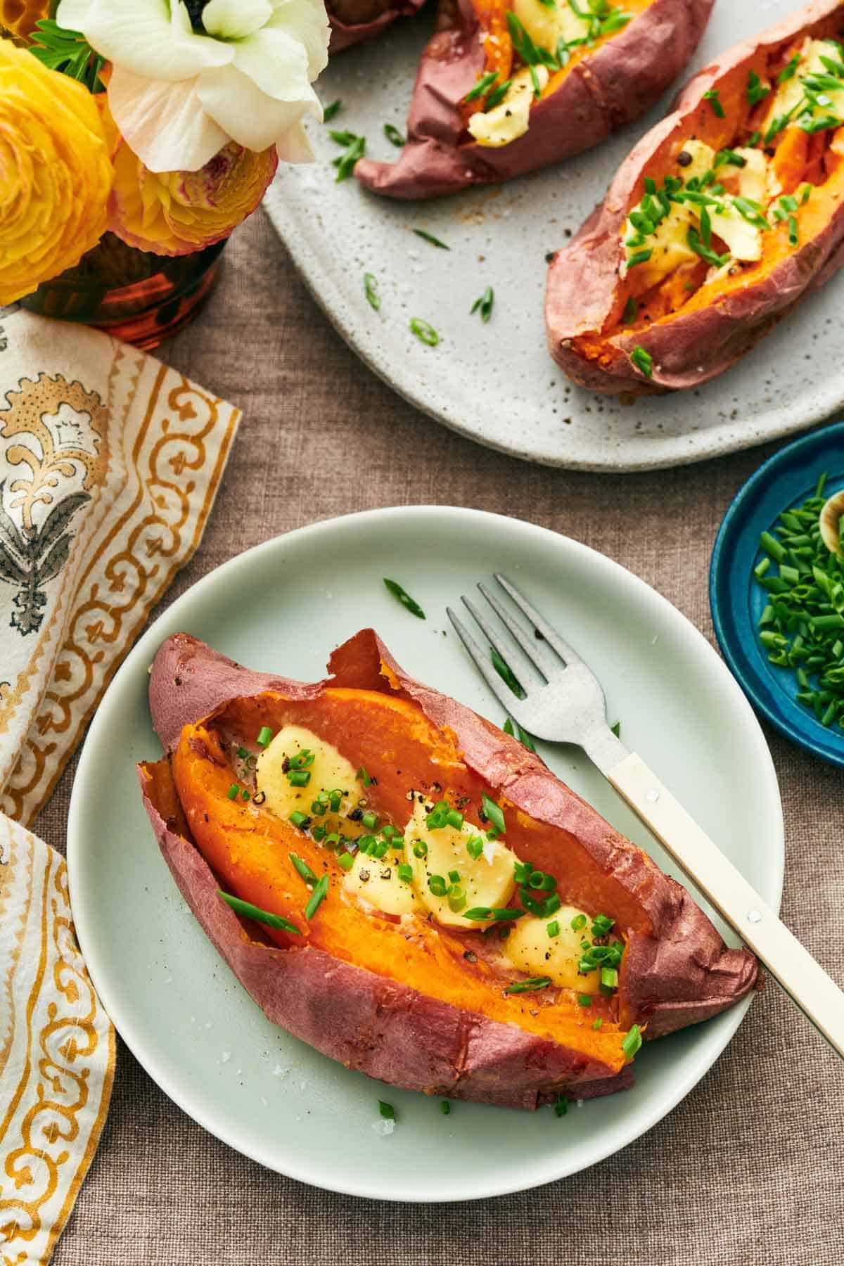 A plate with a baked sweet potato topped with chives and butter. More baked sweet potatoes off to the side along with a bowl of chives and a vase of flowers.