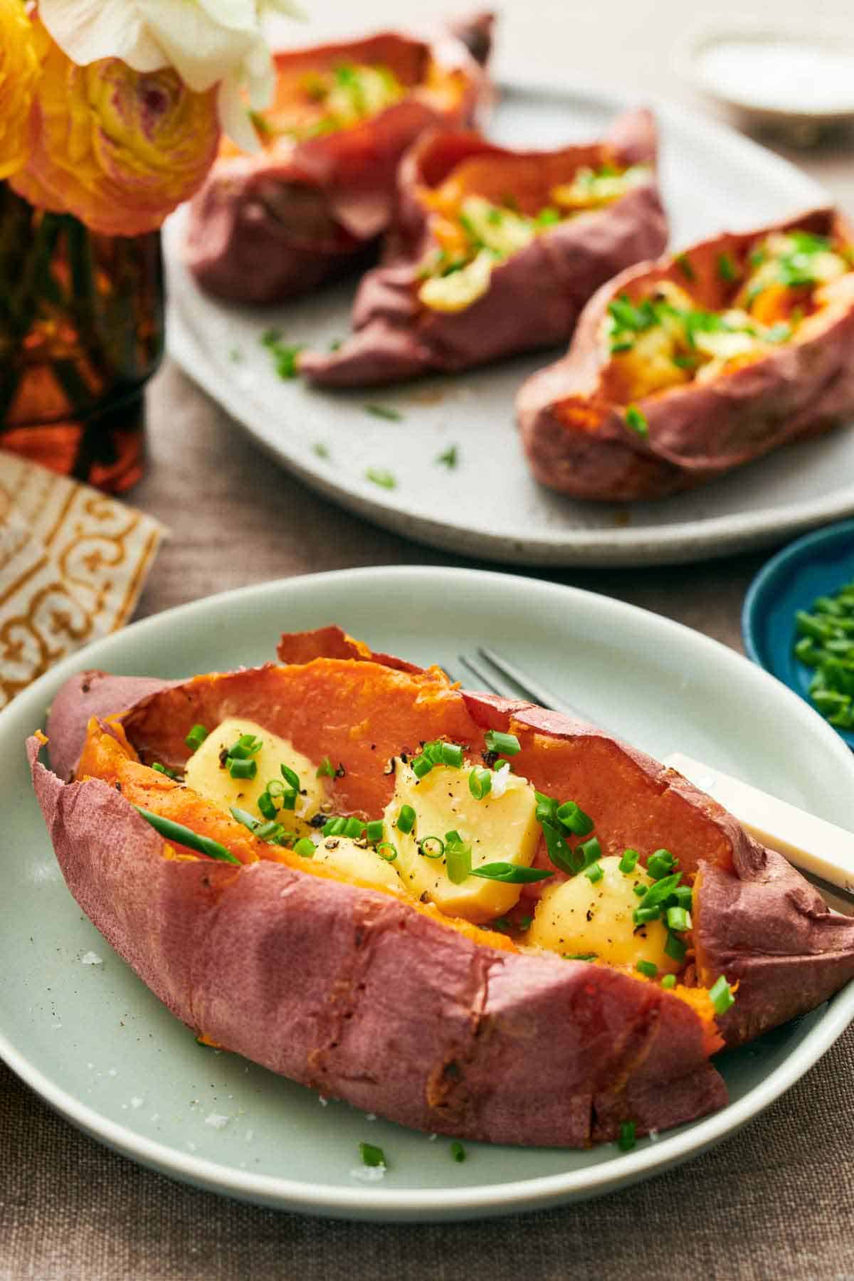 A baked sweet potato garnished with chives and butter on a plate with a fork. In the background is a platter with three more sweet potatoes.