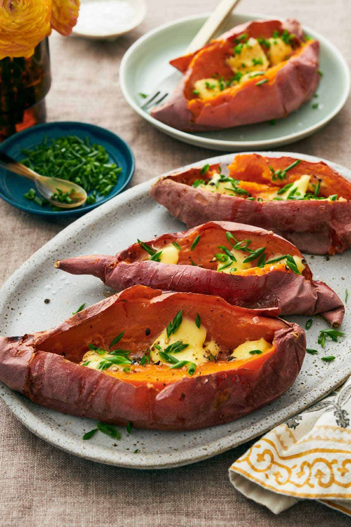 An oval with three baked sweet potatoes with a plated one in the background. A plate of chopped chives off to the side.