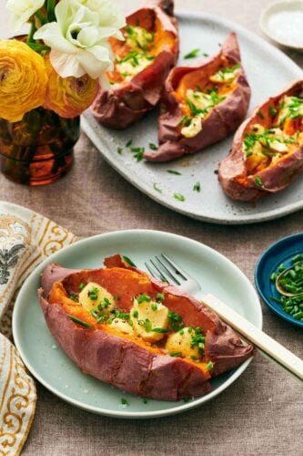 A plate with a baked sweet potato garnished with butter and chives with a fork. A platter with three more in the background along with some flowers in a vase.