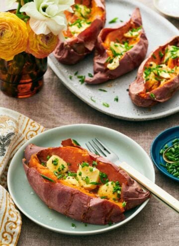 A plate with a baked sweet potato garnished with butter and chives with a fork. A platter with three more in the background along with some flowers in a vase.