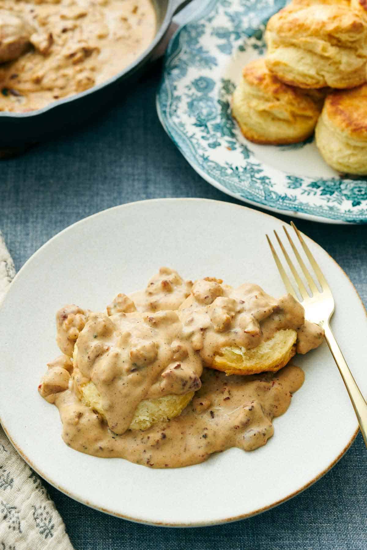 A white plate of biscuits and gravy with a fork. A plate of biscuits in the back along with a skillet.