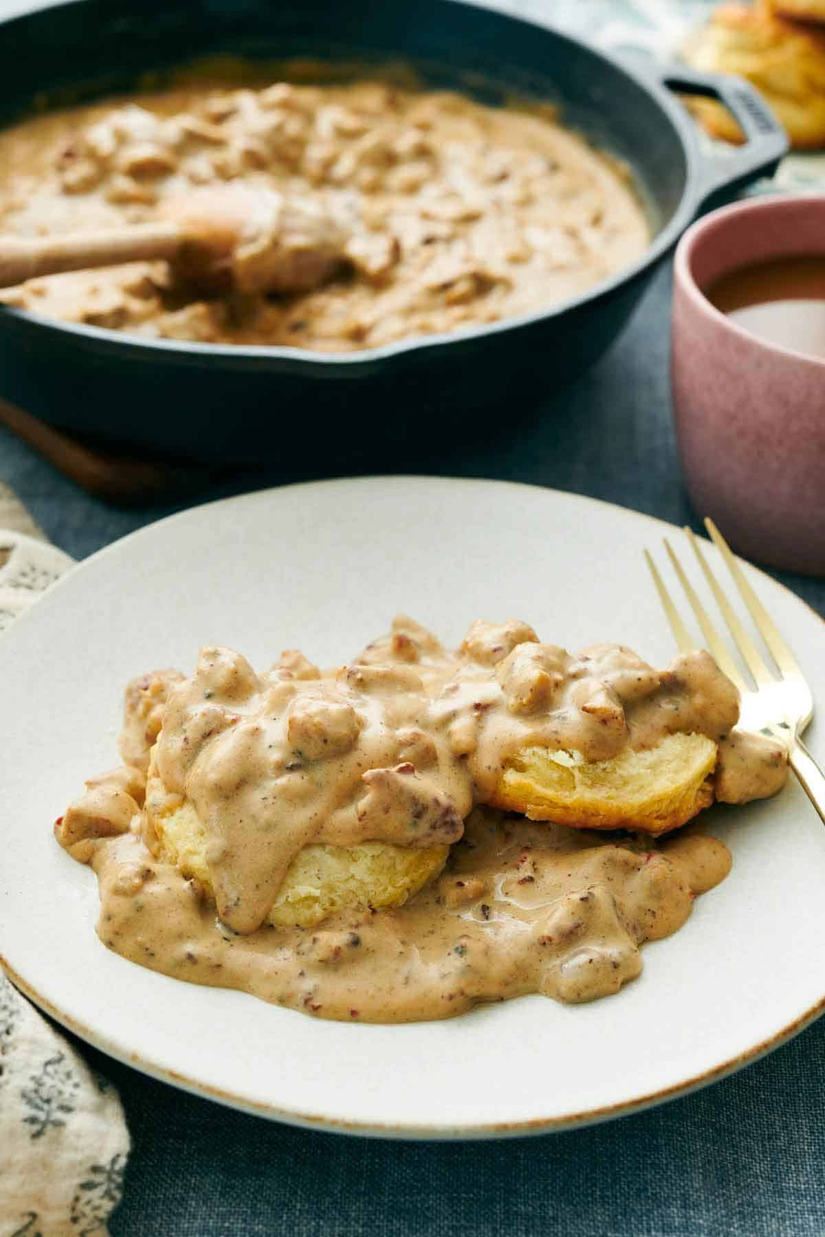 A plate of biscuits and gravy with a skillet of more in the background along with a pink mug.