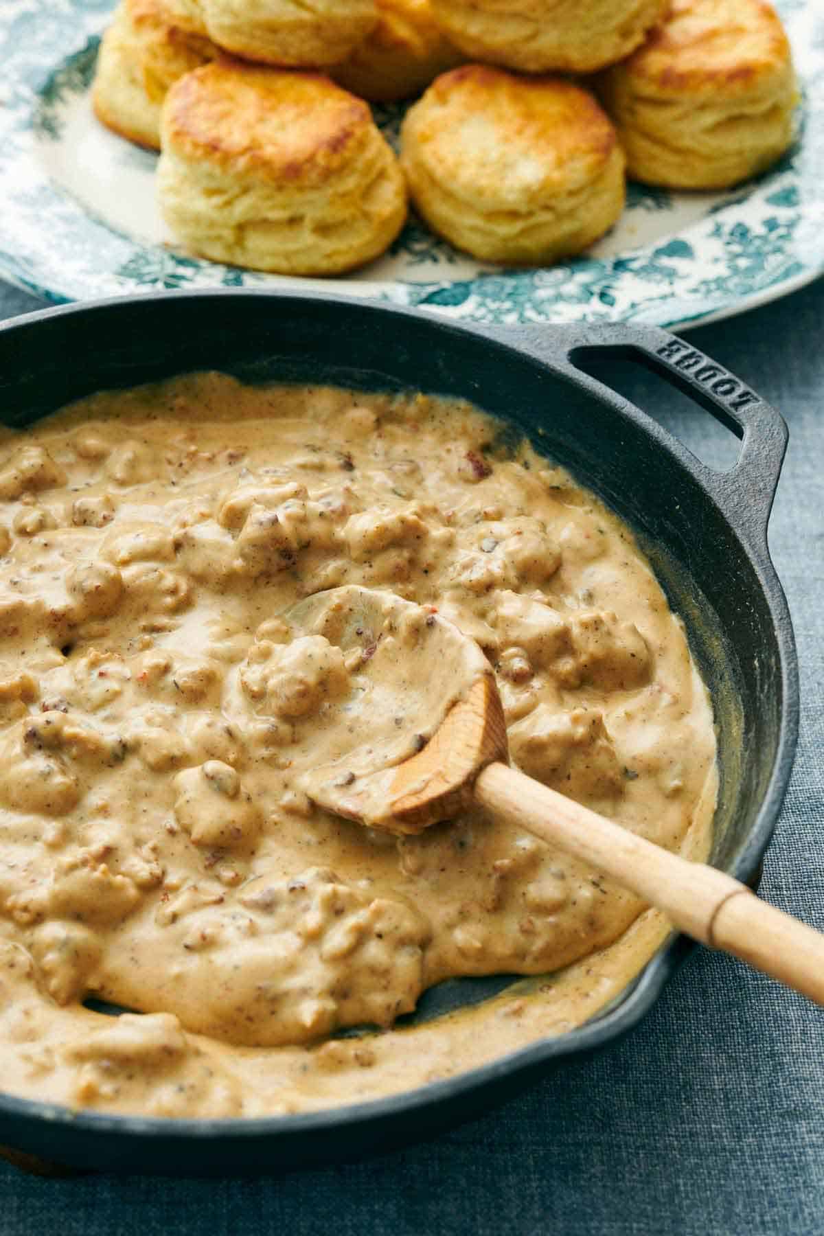 A skillet of biscuits and gravy with a wooden spoon. A platter of biscuits in the background.