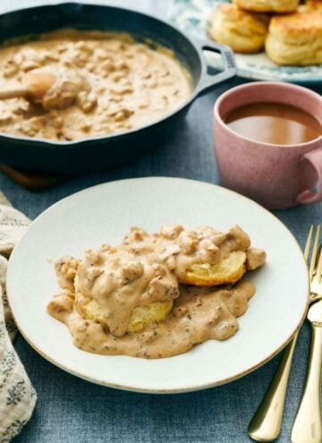 A plate of biscuits and gravy with a skillet of more gravy in the background. A drink, biscuits, and forks behind the plate.