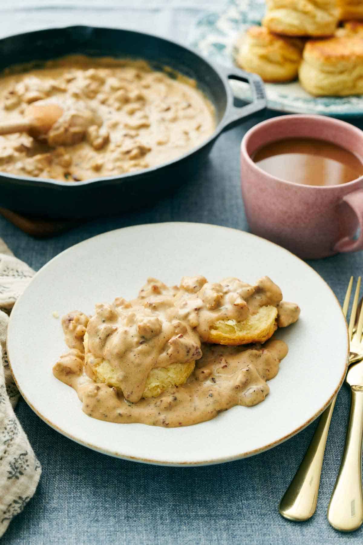 A plate of biscuits and gravy with a skillet of more gravy in the background. A drink, biscuits, and forks behind the plate.