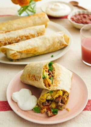 A plate with a breakfast burrito cut in half with sour cream on the side. More breakfast burritos in the background on a platter with sour cream and salsa.