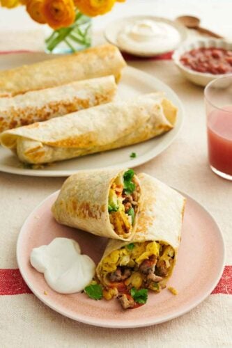 A plate with a breakfast burrito cut in half with sour cream on the side. More breakfast burritos in the background on a platter with sour cream and salsa.