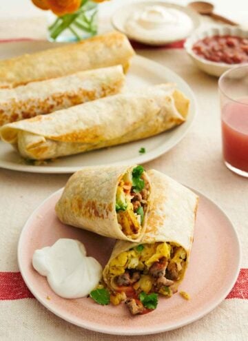 A plate with a breakfast burrito cut in half with sour cream on the side. More breakfast burritos in the background on a platter with sour cream and salsa.