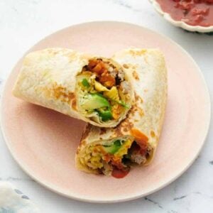 A pink plate with a breakfast burrito, cut in half, with one half leaned on top of the other. A bowl of sour cream and salsa on the side.